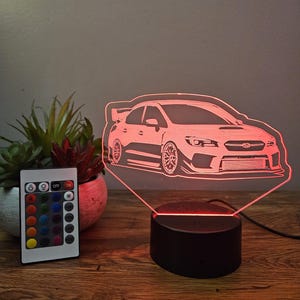 Può includere: Una luce notturna LED 3D a forma di auto bianca con una base nera e un telecomando. Il telecomando ha pulsanti per diversi colori e un pulsante bianco per la luce bianca.
