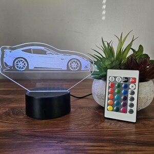 May include: A white acrylic 3D night light shaped like a sports car with a black base and a remote control with 16 color options.