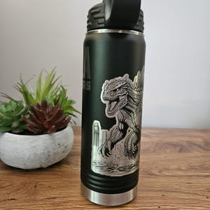 May include: A black and green stainless steel water bottle with a black lid and a silver bottom. The bottle has a graphic of a Godzilla-like monster on the side.