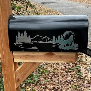 Könnte beinhalten: Schwarzer Briefkasten mit einem Berg- und Wolfsmotiv. Der Briefkasten ist an einem Holzpfosten befestigt.