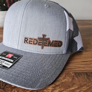 Puede incluir: Una gorra de camionero gris y blanca con un parche de cuero marrón que dice "REDEEMED" con una cruz en la parte superior. La gorra está hecha por Richardson.