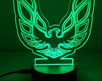 Puede incluir: Una luz nocturna LED verde con forma de logotipo de Pontiac Firebird. El logotipo es un pájaro estilizado con las alas extendidas. La luz está sobre una base negra con un interruptor.