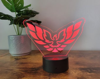 Peut inclure: Une veilleuse LED rouge en forme d'oiseau stylisé avec les ailes déployées. La lumière est sur une base noire.