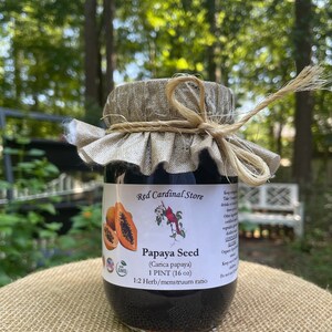 May include: A glass jar with a brown lid and a hessian fabric cover. The jar contains a dark liquid and has a label that reads "Papaya Seed (Carica papaya) 1 PINT (16 oz) 1:2 Herb/menstruum ratio". The label also has a red cardinal logo and a non-GMO certification.