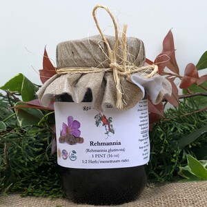 May include: A glass jar with a brown lid and a burlap cloth tied around it. The jar contains a dark liquid and has a label that reads "Rehmannia (Rehmannia glutinosa) 1 PINT (16 oz) 1:2 Herb/menstruum ratio". The label also includes a small image of a red cardinal and a purple flower.