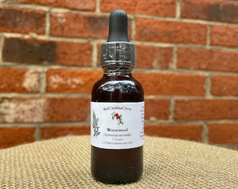 May include: A small, dark brown glass bottle with a dropper top. The bottle is labeled "Red Cardinal Store" and contains "Wormwood (Artemisia anomala)" with a 1:2 herb/menstruum ratio. The bottle is sitting on a woven brown surface.