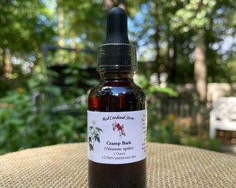 May include: A dark brown glass bottle with a black dropper top. The bottle is labelled "Red Cardinal Store" and "Cramp Bark (Viburnum opulus)" with a 1:2 herb/menstruum ratio. The bottle is sitting on a woven brown surface.