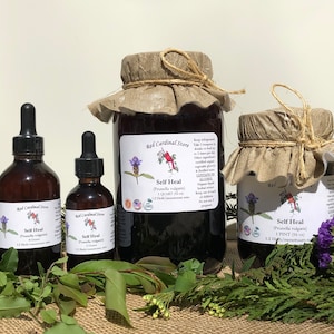 May include: An assortment of amber glass bottles and jars of varying sizes, labeled "Self Heal" from Red Cardinal Store. The containers are sealed with fabric and twine, surrounded by greenery. The labels include botanical illustrations and text.