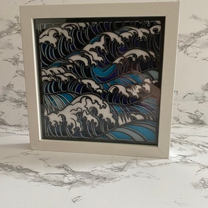 May include: A framed paper art piece depicting a stylized wave pattern in shades of blue, white, and black. The waves are layered and create a sense of depth and movement.