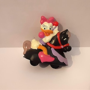 May include: A pink and white duck figurine riding a black horse with purple and red accents. The horse has a red bridle and a purple saddle.
