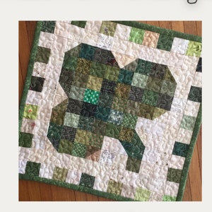 May include: A scrappy mini quilt pattern featuring a shamrock design in shades of green. The quilt is 12 inches by 12 inches and is finished. The pattern is called "Shamrock Shenanigan" and is by Lakegirl Quilts.