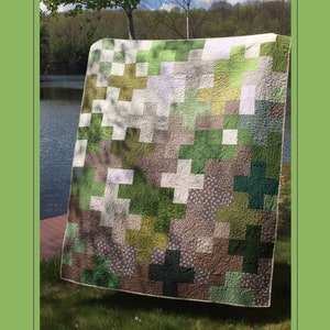 May include: A green, brown and white patchwork quilt with a cross pattern. The quilt is hanging on a clothesline in front of a lake and trees. The quilt is 60 inches by 72 inches finished. The text "lakegirlquilts" is at the bottom of the image.