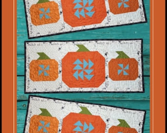 以下が含まれることがあります： lakegirlquiltsによるテーブルランナーの型紙「Pumpkin Picking」、パターン番号046、サイズは35.56cm x 81.28cmです。白い背景に、緑の茎と水色の風車のアクセントが付いた3つのオレンジ色のカボチャがデザインされています。