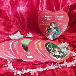 May include: A heart-shaped tarot card box and deck with red card backs. The box features the title "The Heart Tarot" in multiple languages. Clear quartz crystals are arranged near the cards on a red velvet surface.