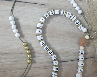 Könnte beinhalten: Eine grüne Schnur mit weißen und goldenen Perlen, die das Alphabet buchstabieren. Die Schnur hat zwei braune, blumenförmige Anhänger.