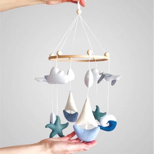 May include: A nautical-themed hanging mobile featuring a circular wooden frame with white strings suspending felt shapes including a sailboat, a seagull, white clouds, teal starfish, and a blue wave, all accented with small wooden beads.