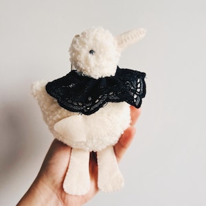 May include: A plush, off-white lamb toy with a black lace collar. The lamb has a fluffy body, felt ears, and felt legs. The toy is held in a hand against a plain white background.