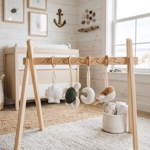 May include: A wooden baby activity gym featuring a frame with carved wave patterns and four hanging plush toys. The toys include a white seashell, a green and white turtle, a white crescent moon, and a brown and white whale, all suspended by ribbons.