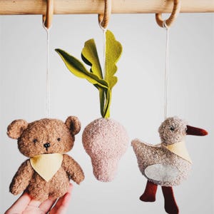 Könnte beinhalten: Ein Baby-Mobile aus Holz mit drei hängenden Plüschtieren: ein brauner Teddybär mit gelbem Lätzchen, eine rosa Radieschen mit grünen Blättern und eine graue Ente mit gelbem Lätzchen und roten Füßen.
