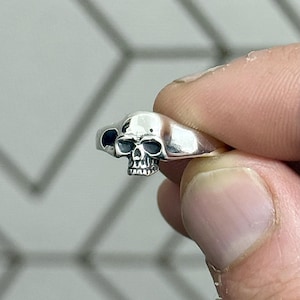 May include: A silver skull ring with detailed facial features. The ring has a polished finish, with the skull's eye sockets and teeth clearly defined. The ring is held up by a person's fingers.