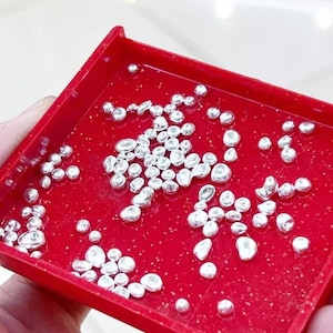 Op de afbeelding: Een rode plastic bak gevuld met talrijke kleine, glanzende, zilverkleurige metalen kralen. De kralen variëren in grootte en vorm en reflecteren licht. De bak wordt door een hand vastgehouden, wat wijst op een context van sieraden maken of knutselen.