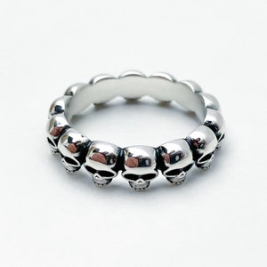 May include: Silver ring with a band of skulls. The ring is made of a smooth, polished metal and features a series of small, detailed skulls that are evenly spaced around the band.