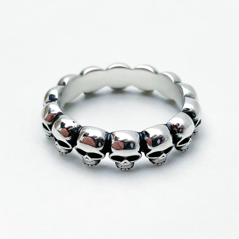 May include: Silver ring with a band of skulls. The ring is made of a smooth, polished metal and features a series of small, detailed skulls that are evenly spaced around the band.