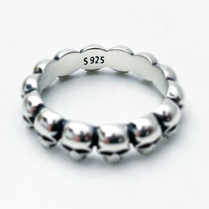 May include: A silver ring featuring a repeating pattern of skull heads. The inside of the ring is marked with "S 925".