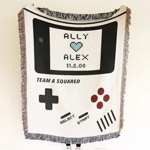 May include: Cream-colored woven blanket with a black and white video game controller design. The blanket features the names "ALLY" and "ALEX" with a pixelated heart and the date "11.2.08". The words "TEAM A SQUARED" are also printed on the blanket.