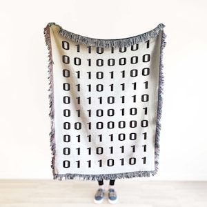May include: A white woven throw blanket with a black binary code pattern. The blanket is hanging from a hook on a white wall.