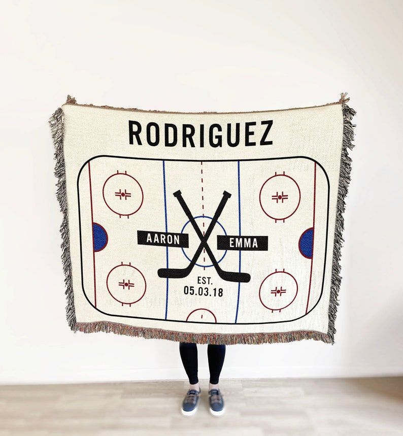 May include: A white woven blanket with a hockey rink design. The rink has two blue circles with red outlines and the text "AARON" and "EMMA" on either side of the center. The text "RODRIGUEZ" is at the top of the blanket and "EST. 05.03.18" is at the bottom.