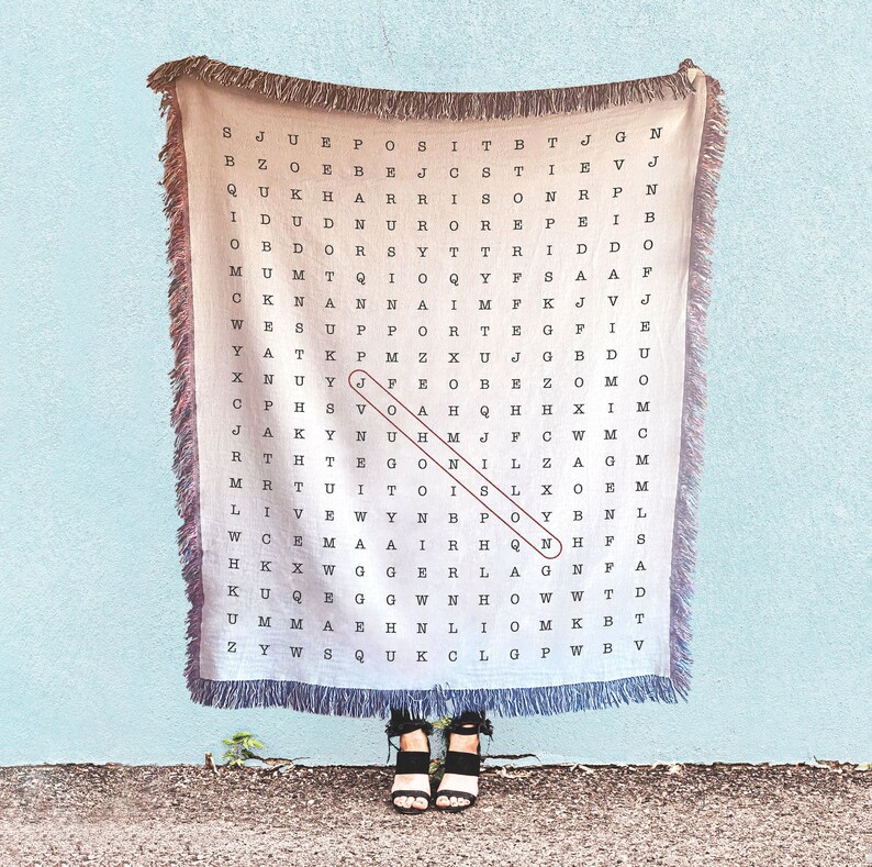 May include: A beige woven tapestry with a black and white word search puzzle. The puzzle features the word "JOY" highlighted in red. The tapestry is hanging on a light blue wall.