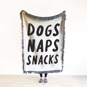 May include: A white woven throw blanket with black text that reads "Dogs Naps Snacks".