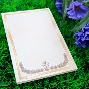 May include: A cream-coloured rectangular notepad with a gold border. The bottom of the notepad features a gold laurel wreath design with a crown emblem. The notepad is resting on green grass, with purple flowers in the background.