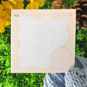 May include: Square notepad with a light beige border and a white interior. The notepad features a cartoon chick illustration in the bottom right corner and a small egg illustration in the top left corner. The notepad is surrounded by yellow flowers and green grass.