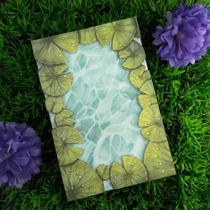 May include: A rectangular card with a watercolour design of a pond and lily pads. The lily pads are green with water droplets, framing a light blue and white water pattern. Two purple flowers and green grass surround the card.