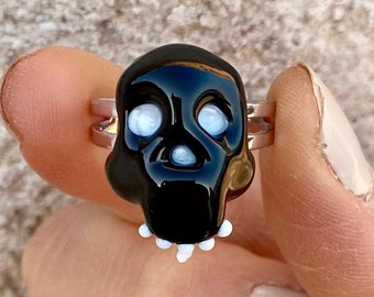 Puede incluir: Un anillo de calavera negro con ojos y dientes blancos. El anillo está hecho de vidrio y está montado en una banda plateada.