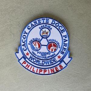 Puede incluir: Una taza de cerámica blanca con un asa negra y el texto "But First, Coffee" en un estilo manuscrito. La taza está sobre una superficie blanca. La taza mide aproximadamente 10 cm de alto.