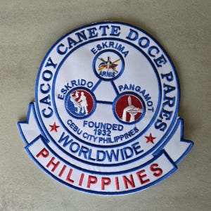 May include: Embroidered patch with a circular design featuring text and graphics. The patch includes the words "Cacoy Canete Doce Pares" and "Philippines" in red and blue. It also has the text "Eskrima," "Eskrido," and "Pangamot."