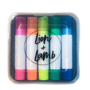 May include: A set of six colorful markers in a clear plastic case. The markers are arranged in a rainbow pattern, with the colors pink, orange, yellow, green, blue, and purple. The case has a black circle with the text "Lion + Lamb" printed on it.