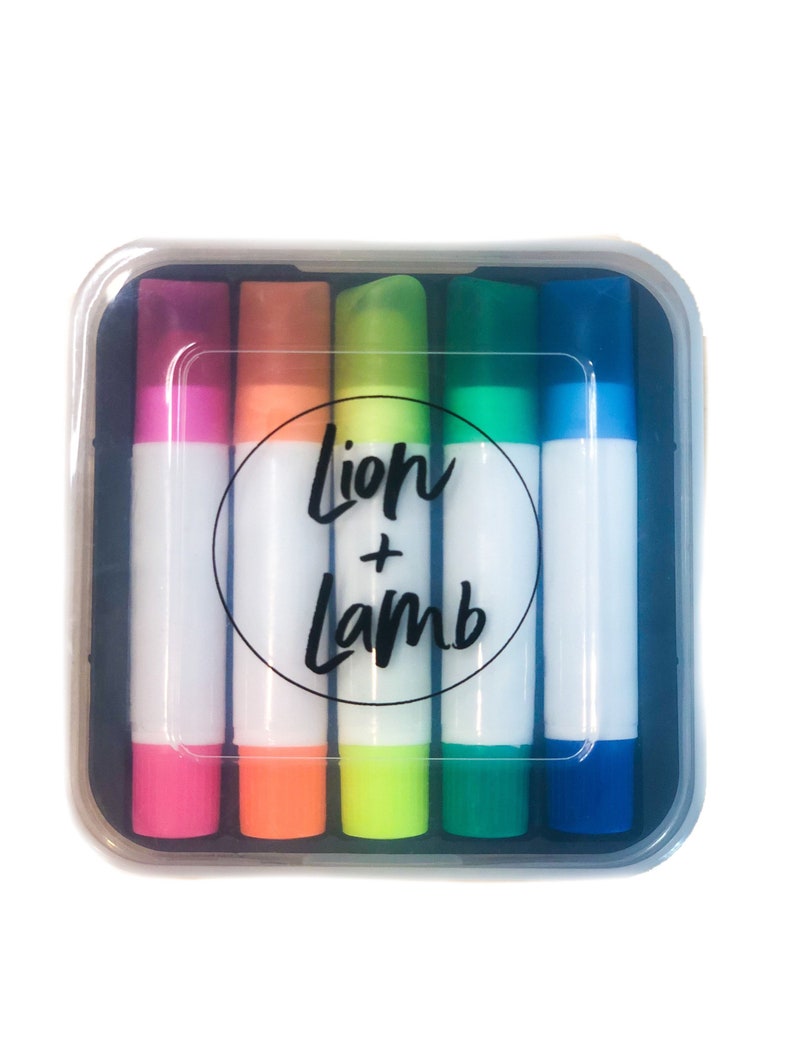 May include: A set of six colorful markers in a clear plastic case. The markers are arranged in a rainbow pattern, with the colors pink, orange, yellow, green, blue, and purple. The case has a black circle with the text "Lion + Lamb" printed on it.