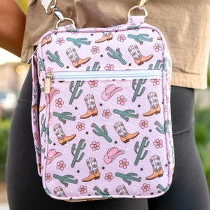 Pode incluir: Uma bolsa rosa de ombro com um zíper branco e uma estampa de cacto e bota de cowboy.