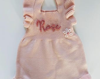 May include: A pink knitted baby onesie with ruffled sleeves and a flower embroidered on the front. The onesie is personalized with the name "Rose" embroidered in pink thread.