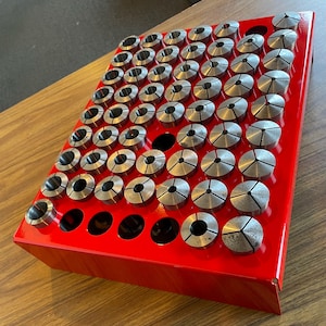 May include: A red metal storage tray with a grid of circular holes, filled with a collection of silver metal collet chucks.