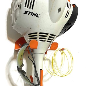 May include: A white and orange STIHL string trimmer is mounted on a wall-mounted storage rack. The trimmer has a black handle and a yellow cord wrapped around the rack. Safety glasses are also hanging on the rack.