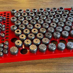 May include: A red metal storage tray with a grid of circular holes, filled with a variety of silver collet chucks. The chucks are arranged in rows and columns, with the largest chucks at the top and the smallest chucks at the bottom.