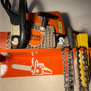 Op de afbeelding: Oranje kettingzaag wandbevestiging met een Stihl MS 250C kettingzaag die eraan hangt. De bevestiging heeft een kettingzaag silhouet en haken om kettingen op te hangen.