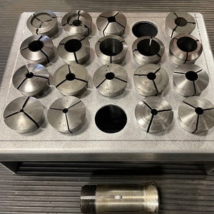May include: A collection of metal collets, each with a split design, arranged in a rectangular metal holder. The collets are various sizes and shapes. A single collet is positioned below the holder. The items appear to be used for holding workpieces in a lathe or similar machine.