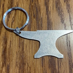 May include: A silver metal keychain shaped like an anvil. The keychain has a silver ring attached to the top.