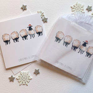 Cartes de Noël de luxe. Aquarelle de moutons mangeant de l'herbe avec un autre vers le haut portant un bonnet de Noel. Orné d'une pierre précieuse scintillante.